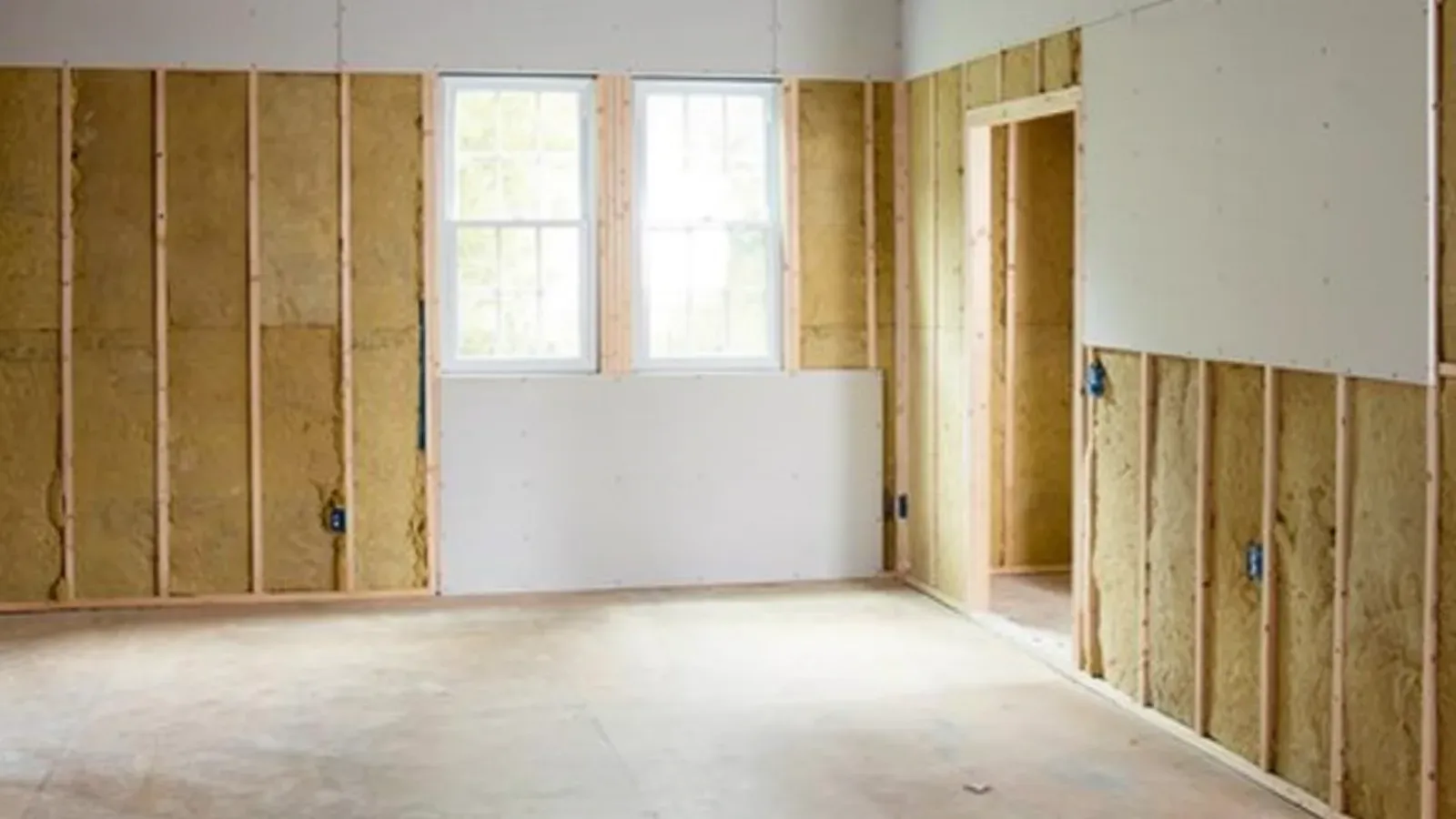 An empty framed room mid-installation, one wall sheeted with bare gypsum drywall while the remaining walls are still open studs with insulation
