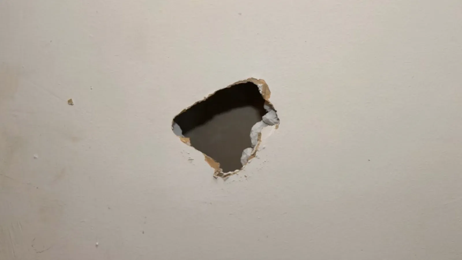 Small ragged hole broken through a painted interior drywall wall, torn paper and crumbling gypsum around its edges exposing the dark cavity behind