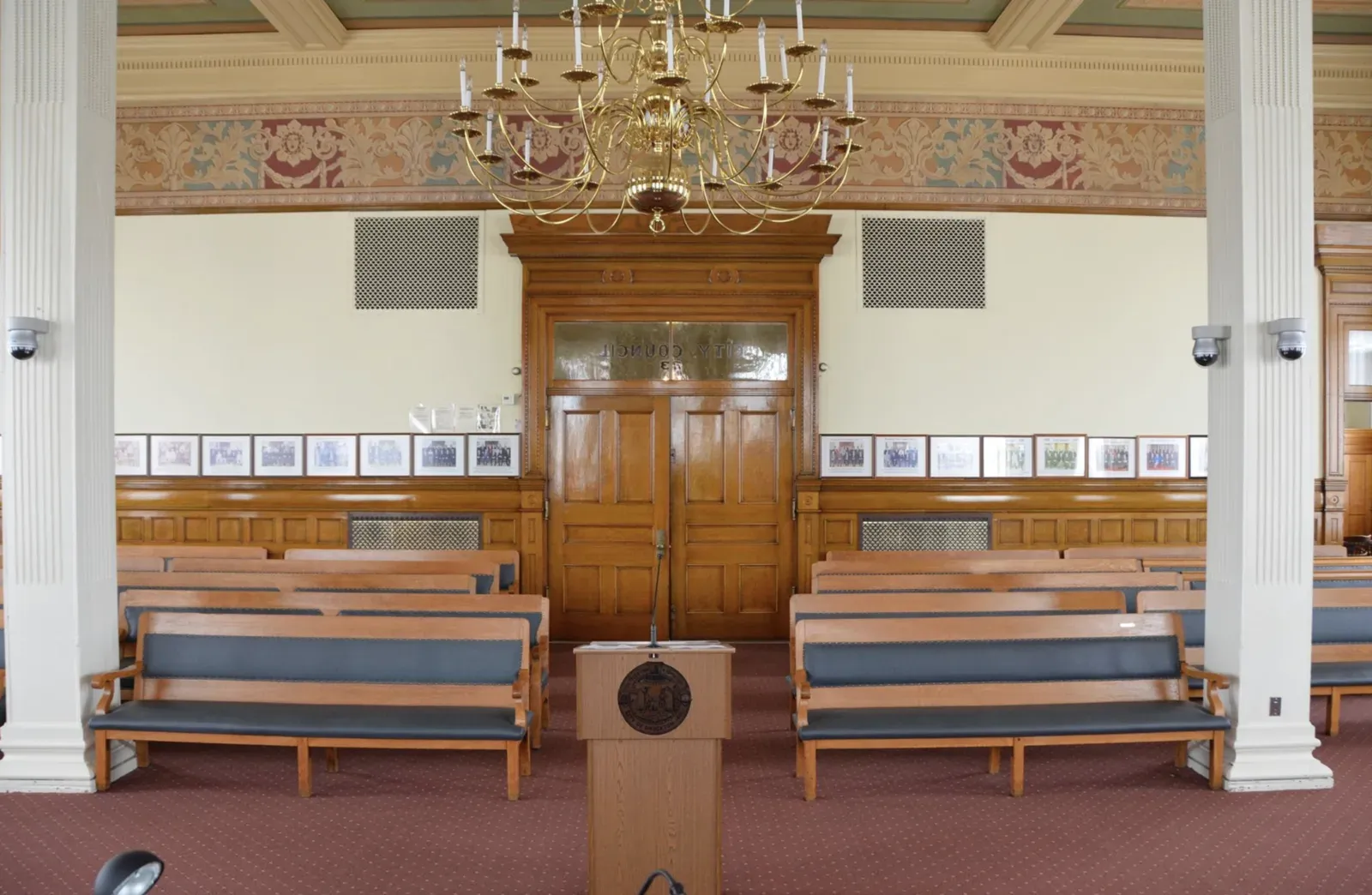 Brockton City Hall council chamber with a stenciled plaster frieze, a brass chandelier, oak-panelled wainscot and doors, and plaster-finished walls above rows of wooden benches