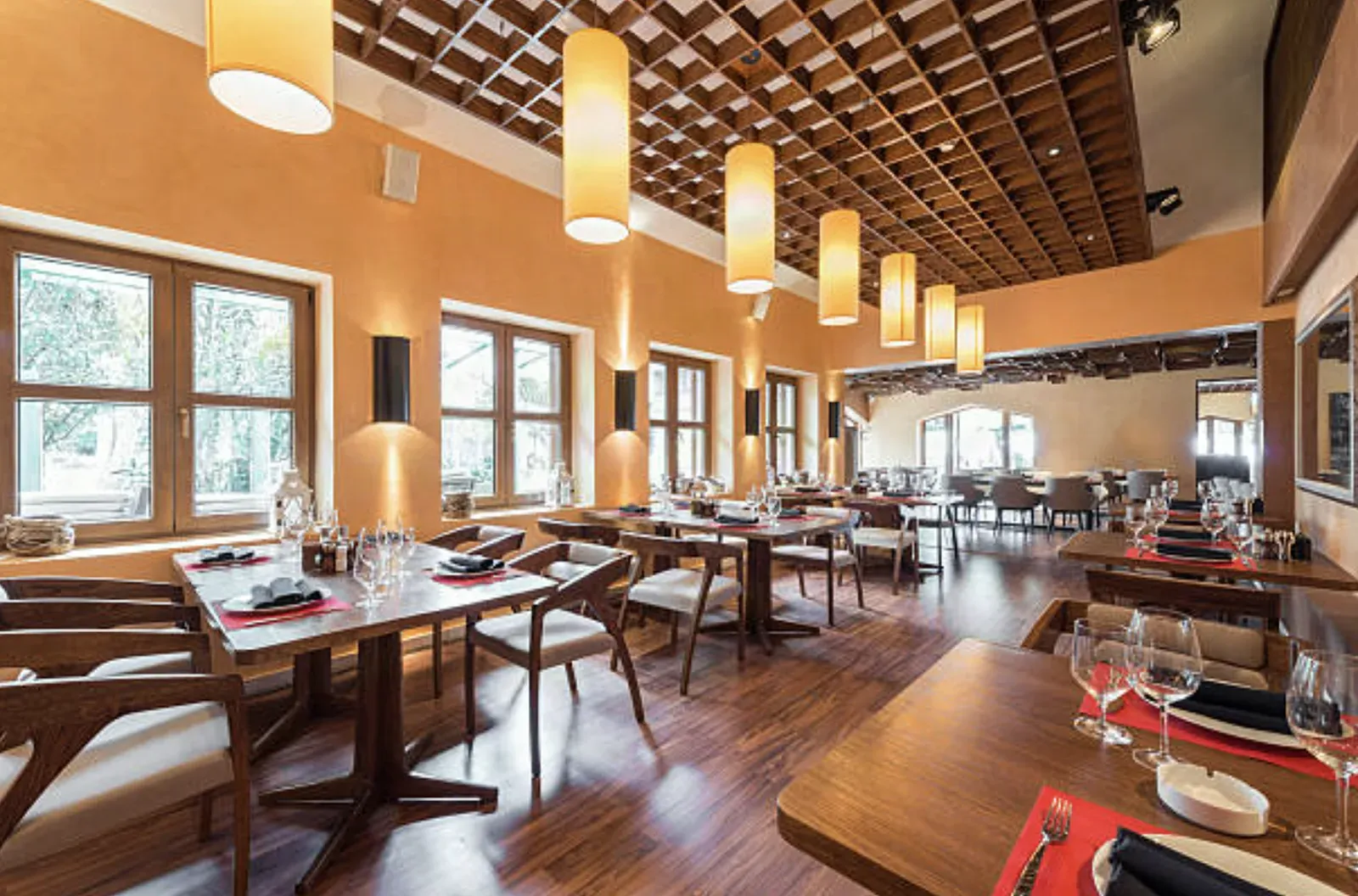Restaurant dining room with warm plaster-finished walls, a coffered wood-grid ceiling with cylinder pendant lights, dark wall sconces, and tables set beside tall windows